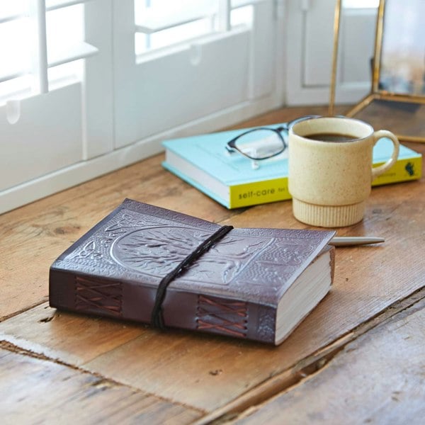 The Tree of Life Leather Journal rests on a wooden table beside a coffee mug, glasses, and a pen. Nearby lies "self-care," its light blue cover adding calm under the window's shuttered light.