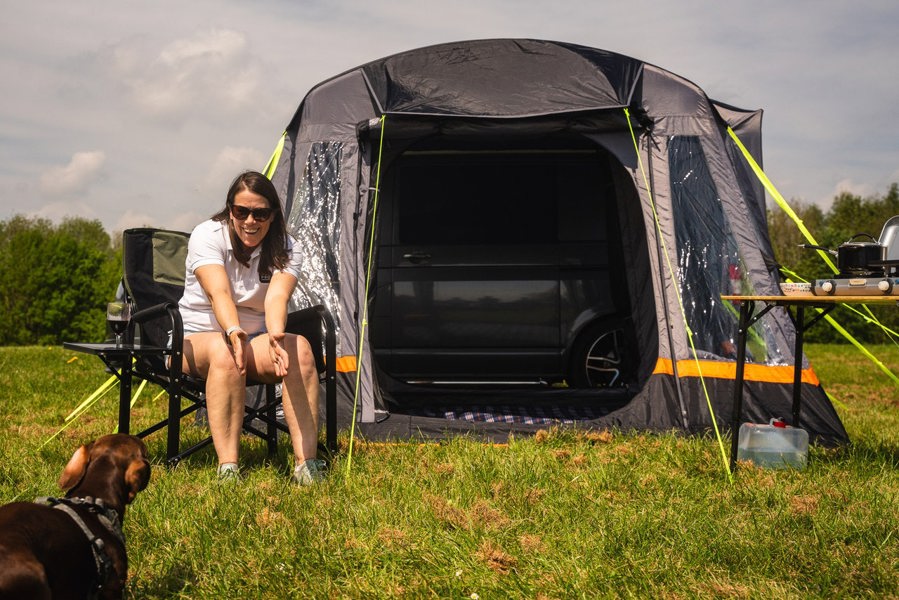 A woman sits in a camping chair outside the Loopo Breeze® v2 Campervan Awning, she is playing with a dog imageset:Charcoal