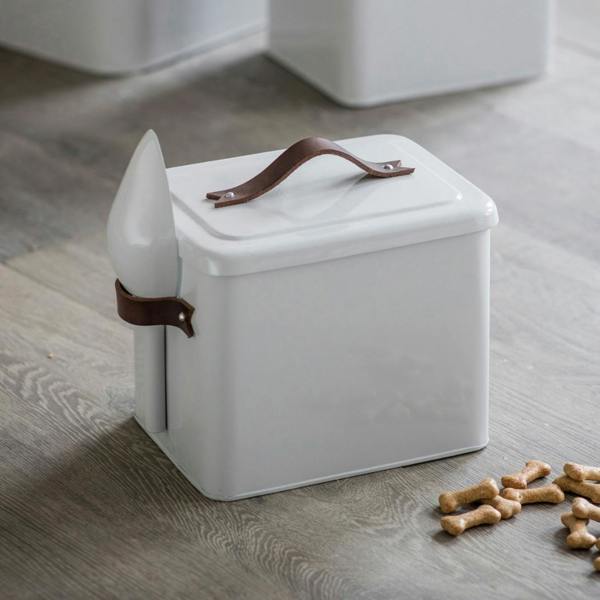 A small white chalk-coloured powder-coated steel pet food bin featuring a brown leather handle on the lid, a matching leather side loop holding a scoop, and bone-shaped treats scattered on the floor.