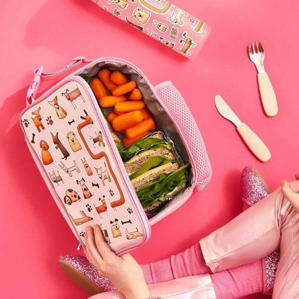 Lunchbox with cartoon dogs holds carrots and sandwiches inside vibrant pink setting with utensils and child wearing pink sparkly shoes.