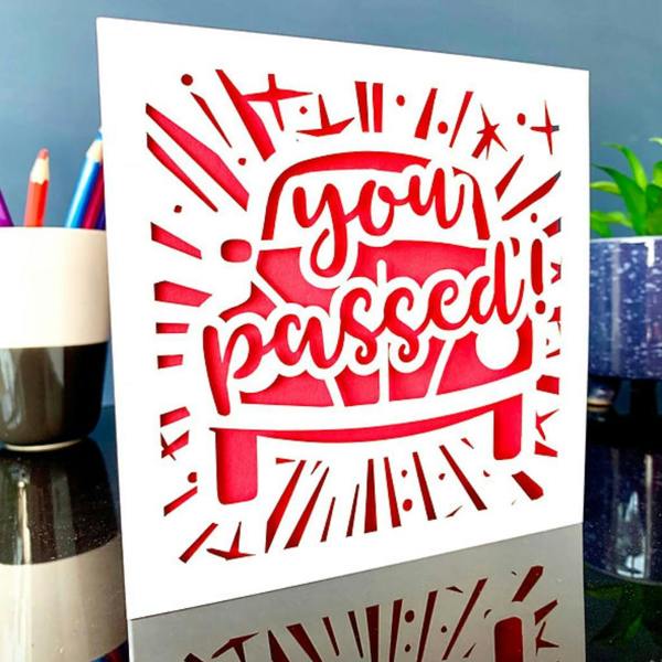 Handmade white papercut driving test card featuring You Passed lettering over a car silhouette with a red paper insert.