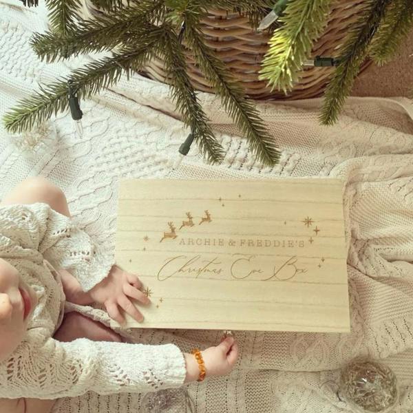 A top-down view of a light wooden Christmas Eve box engraved with the names Archie and Freddie's, featuring flying reindeer and stars, placed on a white cable knit blanket under a Christmas tree next to a young child.