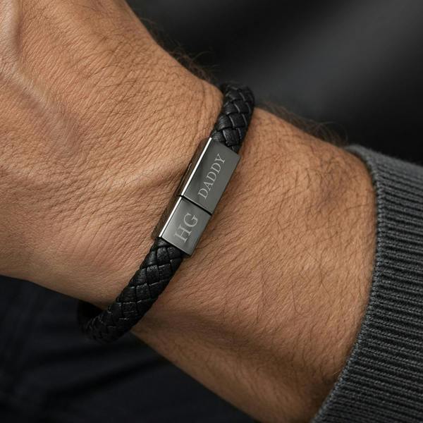 A close-up view of a man's wrist wearing the braided black leather charger bracelet with the dark metallic clasp engraved with HG and DADDY.