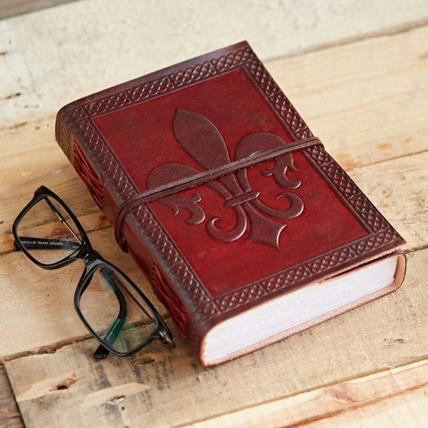 A Fleur de Lis Leather Journal with a red design and wraparound strap lies on a wooden surface beside eyeglasses.