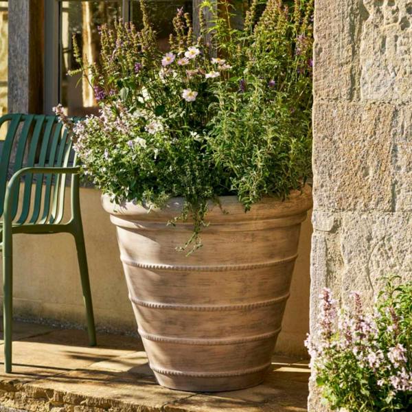 Large 70cm Cotswold ribbed terracotta planter sitting as a standalone feature on a flagstone patio, filled with a massive, overflowing arrangement of purple salvia and pink garden flowers.