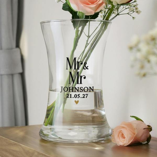 A clear, waisted glass vase sitting on a wooden surface next to a single pink rose. The vase is engraved with the text "Mr & Mr", "JOHNSON", and the date "21.05.27" with a small gold heart beneath.