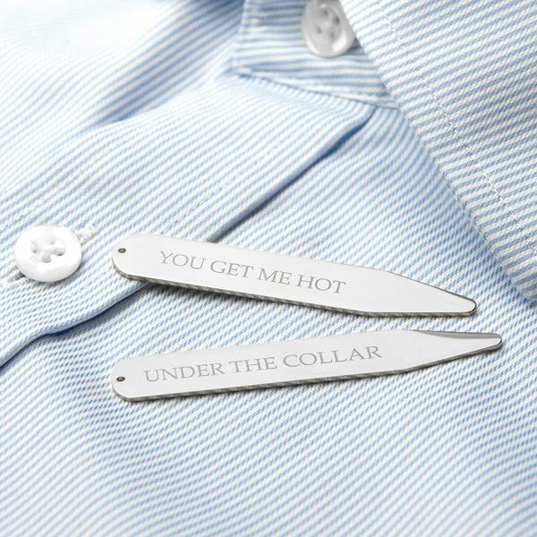 A close-up studio shot of two silver rhodium-plated collar stiffeners resting on a crisp, light blue and white pinstriped men's dress shirt. The stiffener at the top is engraved with the text "YOU GET ME HOT" and the one below it reads "UNDER THE COLLAR". The shirt has a white button visible in the background.