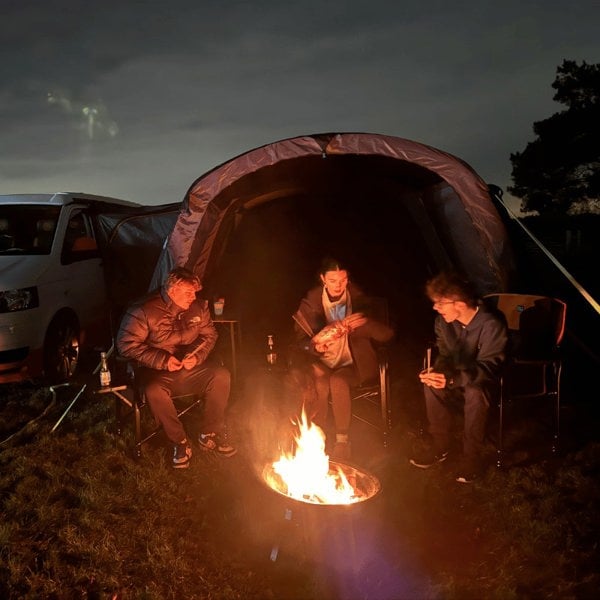 A family sits outside the Cocoon Breeze® v2 Campervan Awning wrapped in blankos surrounding a fire. imageset:Charcoal