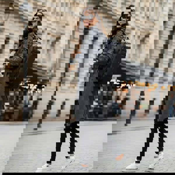 Woman walking on a city street holding a coffee cup and wearing a gray coat with a black commuter laptop best professional backpack by Riseicon.