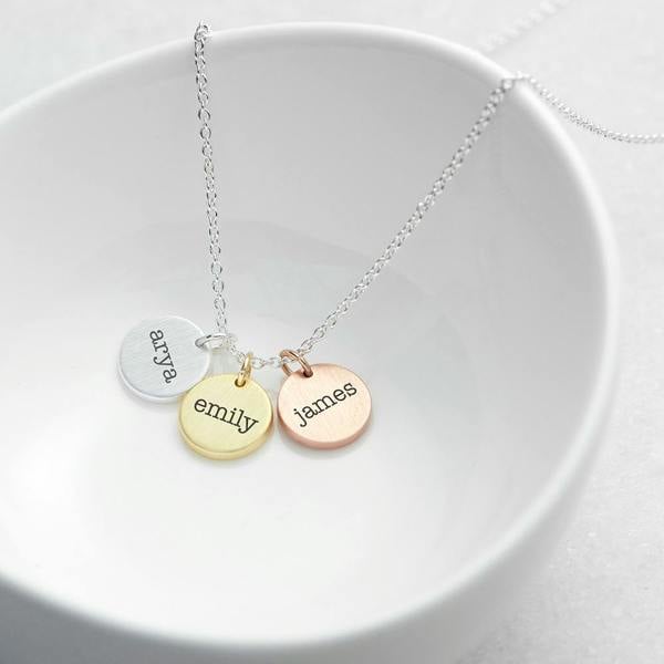 A personalised mixed-metal three-disc necklace featuring lowercase script name engravings for 'arya' on silver, 'emily' on yellow gold, and 'james' on rose gold, beautifully draped inside a small white ceramic dish.