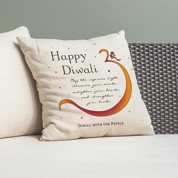 A cream textured linen cushion cover printed with a vibrant orange Om symbol, festive text, and 'Diwali with the Patels' written at the base, resting on a white sofa.