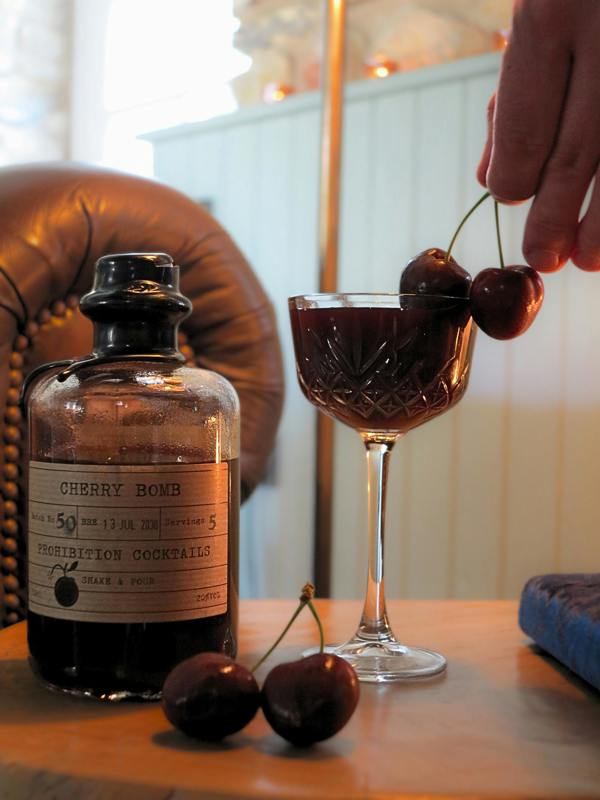 A bottle labeled Cherry Bomb Prohibition Cocktails sits on a wooden table beside a coupe glass filled with a dark red cocktail, with cherries on the glass rim and two cherries on the table. A hand is placing a cherry onto the glass, and a leather chair and indoor setting are visible in the background.