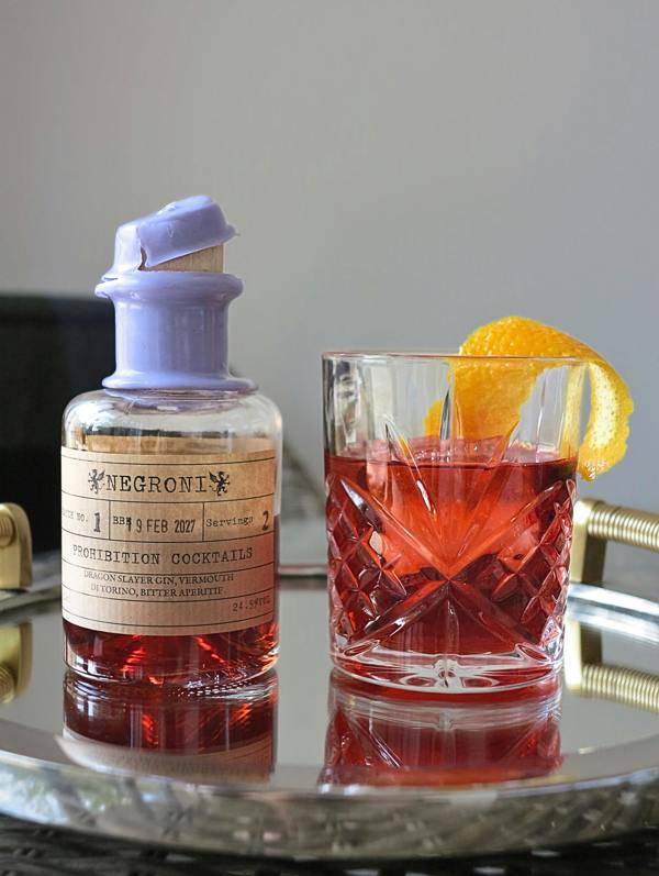 A bottle of Negroni Prohibition Cocktails with a lavender stopper and beige label, standing next to a cut-glass tumbler filled with a red cocktail and garnished with an orange slice, set on a reflective silver tray.