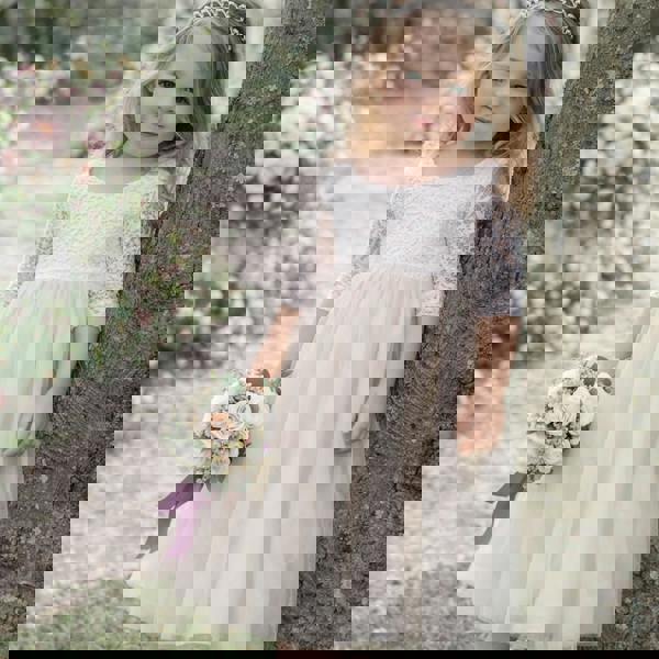 Young girl in a white lace dress holding flowers, standing next to a tree in a floral garden.