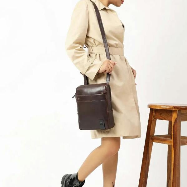 Person wearing a beige trench coat holding a brown leather messenger bag on a white background