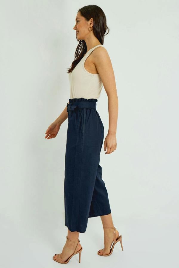 Woman wearing a white sleeveless top and navy paperbag pants on a white background