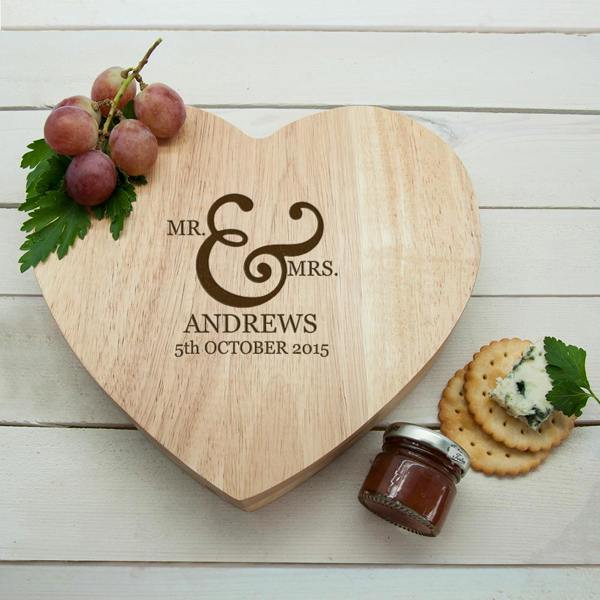 A top-down view of a heart-shaped wooden cheese board engraved with "Mr. & Mrs. Andrews" and a wedding date, styled on a white wooden table next to red grapes, water biscuits, and a small jar of chutney.