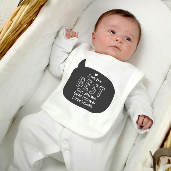 A newborn baby wearing a white personalised cotton bib printed with a black speech bubble that reads 'I am the BEST Gift You Will Ever receive! Love William', lying inside a wicker moses basket.