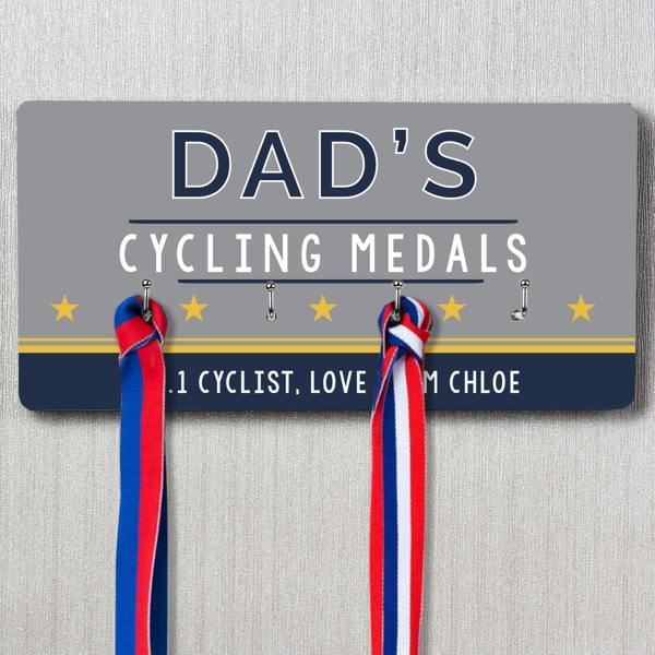 A grey and navy rectangular personalised medal rack hanging on a wall, featuring the text "DAD'S CYCLING MEDALS," five yellow stars, and a personalised message below reading "OUR #1 CYCLIST, LOVE CHLOE." Two medals with red, white, and blue ribbons are shown hanging from the hooks.