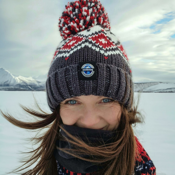 The snow of winter provide the backdrop for woman who is wearing a chunky Nordic knit Super Bobble hat in dark grey with white and red Tyrol design. The hat also has a deep ribbed headband, contains reflective yarns and has a luxury sherpa fleece lining.