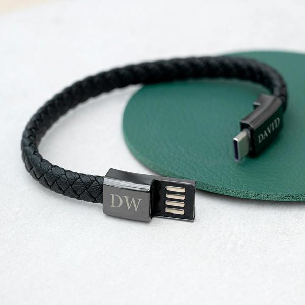 An unclasped braided black leather men's bracelet showing the exposed dark metal USB-A connector plug engraved with DW and the USB-C pin plug engraved with DAVID.
