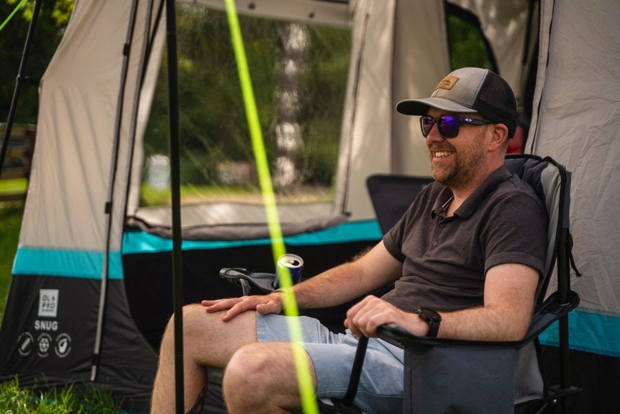 A man sits outside the Snug Poled Tailgate Awning, in an OLPRO camping chair