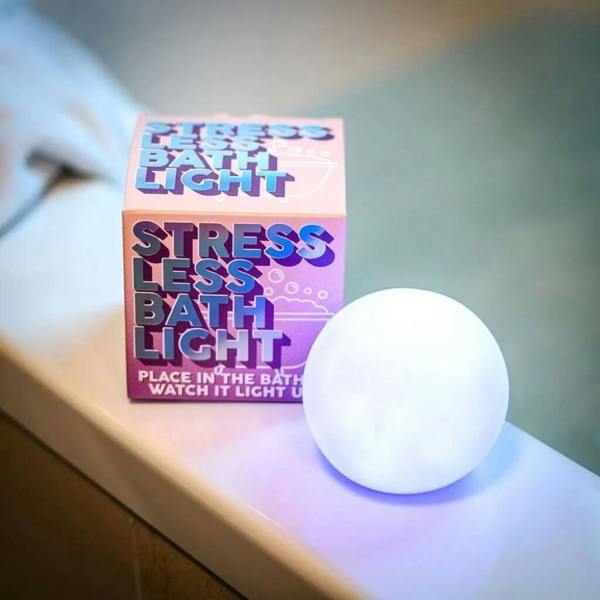 A glowing white spherical bath light illuminating the edge of a clean white bathtub next to its pink and blue retro-style gift box.