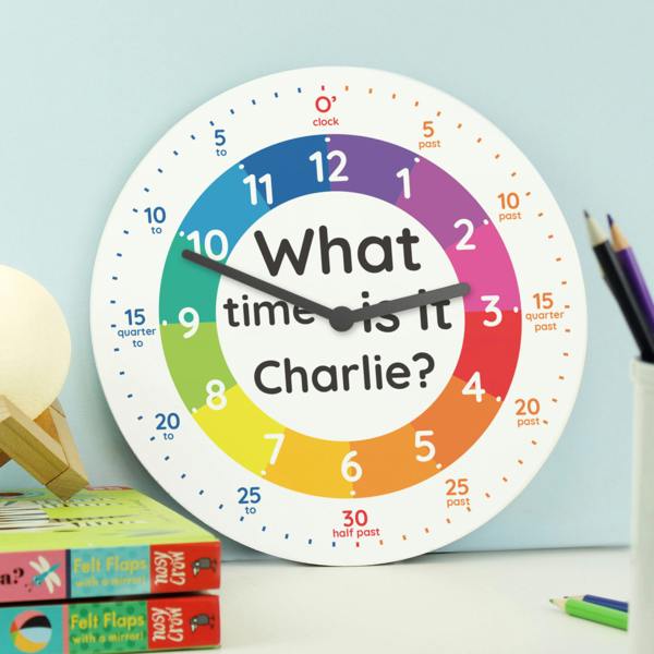 A personalised children's wooden wall clock by Abode Home & Gifts featuring a colourful rainbow time-teaching dial that reads "What time is it Charlie?", styled on a desk next to children's books and a jar of colouring pencils.