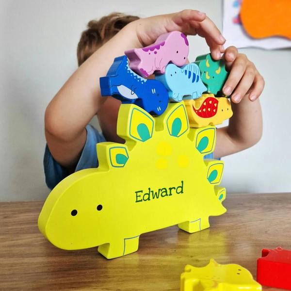 A personalised wooden dinosaur stacking toy featuring a large yellow stegosaurus printed with 'Edward', being built up with smaller colourful wooden dinosaurs by a child on a wooden table.