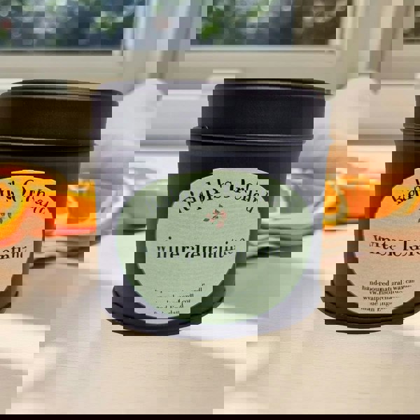 Candle in a black tin labeled 'Ralph's Orchard winter clementine' on a surface with oranges.