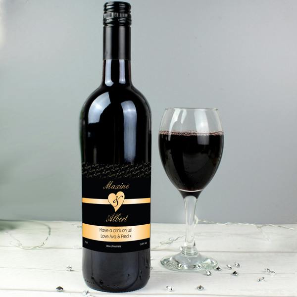 A bottle of Personalised Couples Heart Shiraz Red Wine with a black and gold label next to a filled wine glass on a white wooden surface.