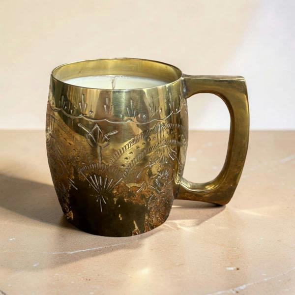 A vintage-style brass mug candle with a textured, engraved floral pattern and a large handle, filled with white wax and sitting on a beige surface.
