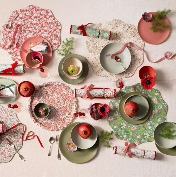 Styled Christmas dinner table setting featuring Liberty print placemats, ceramic plates, pomegranates, flowers, and Liberty print crackers with velvet bows.