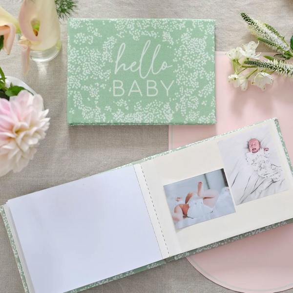 Sage green fabric Hello Baby photo album lying flat and open next to fresh dahlia flowers showing newborn baby pictures.