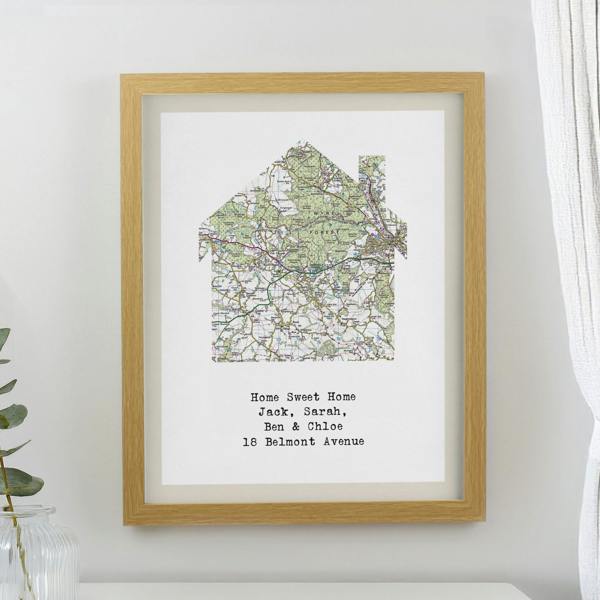 An oak framed print showcasing a colourful Ordnance Survey map cut into a house silhouette, personalised with four lines of custom text, hanging on a neutral wall.
