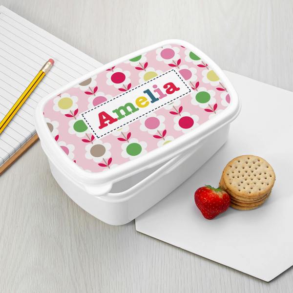 A white plastic personalised girl's lunch box printed with pink sunflower patterns and the name Amelia, placed on a table next to a notebook, pencil, strawberry, and crackers.