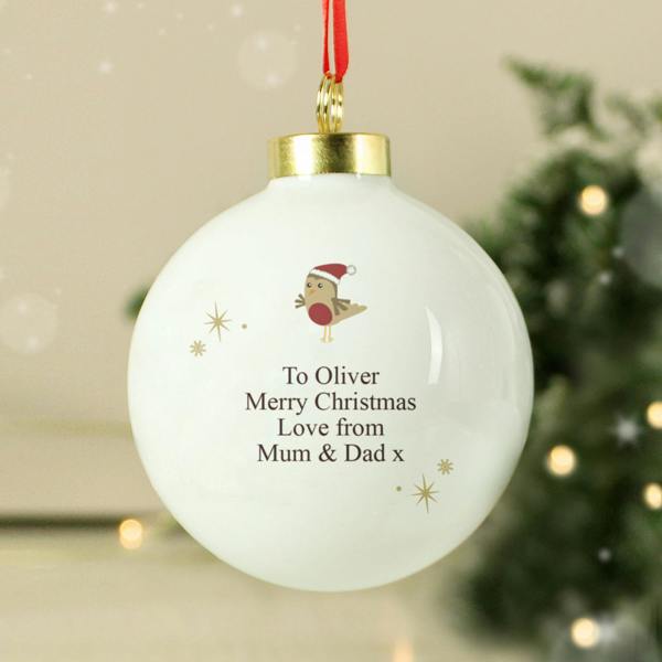 The reverse side of a glossy white ceramic Christmas bauble hanging from a red ribbon. The back features a small illustration of a robin in a Santa hat above a personalized printed message that reads 'To Oliver Merry Christmas Love from Mum & Dad x' surrounded by small gold star accents, with a blurred festive background.