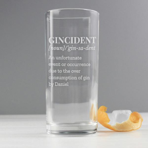 An empty clear highball glass by Abode Home & Gifts featuring a frosted laser-engraved dictionary definition of "Gincident" ending with the name "Daniel", displayed next to a twisted orange peel and a single ice cube on a white tabletop against a grey studio background.