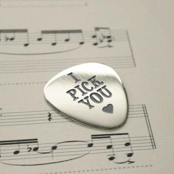A polished steel guitar pick engraved with 'I PICK YOU' and a small heart, resting on a sheet of musical notes.