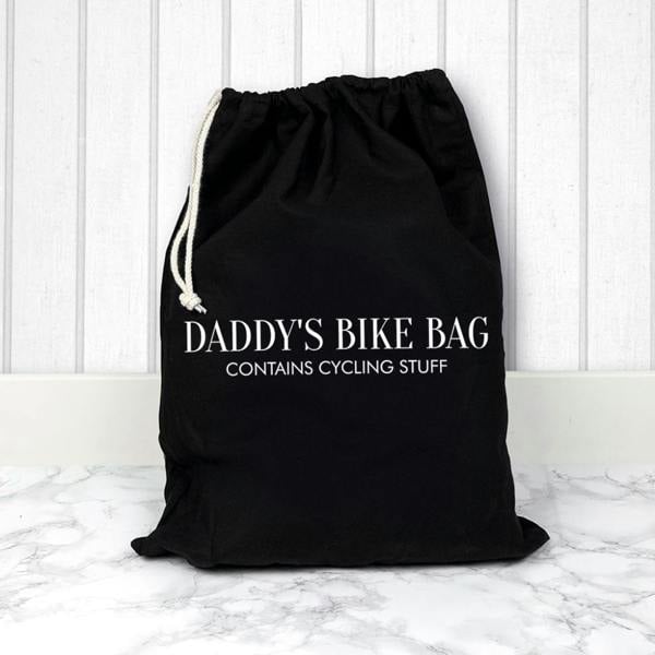 A black cotton drawstring bag printed with DADDY'S BIKE BAG CONTAINS CYCLING STUFF in clean white uppercase typography, sitting on a white marble surface.