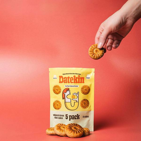 Hand holding a cake above a 'Datekin' 5-pack wheat and bran date cakes bag on a red background