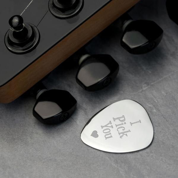 A polished silver guitar pick engraved with 'I Pick You' and a small heart, resting on a grey surface next to the black tuning pegs of a guitar headstock.