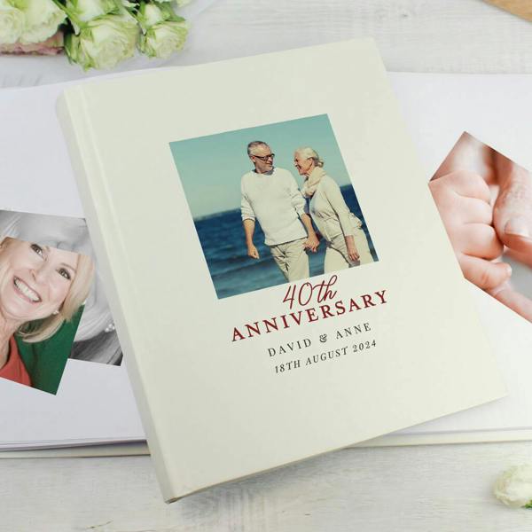 A cream-coloured 40th anniversary photo album lying on a white wooden surface, featuring a photo of an elderly couple walking on a beach on the cover, with the text '40th Anniversary' followed by 'DAVID & ANNE' and '18TH AUGUST 2024'.