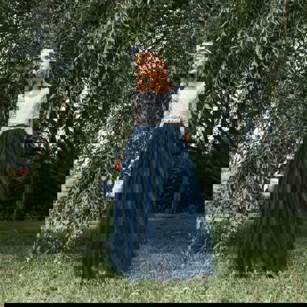 Young girl in a white top and navy blue skirt standing in a grassy area with trees.