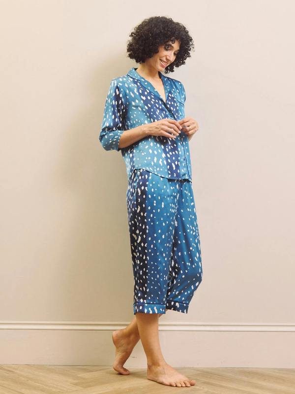 A woman modelling a matching two-piece pyjama set with a blue and white spotted pattern, standing barefoot against a plain beige wall.