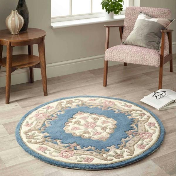 Round decorative rug with floral pattern on a wooden floor, surrounded by a chair and small table.