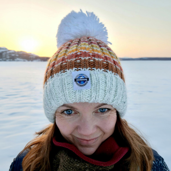 Against a snowy backdrop women wears a striped supeer bobble hat with a base colour of off white and stripes in shades of brown, burnt orange, orange and mustard. The hat also has a deep ribbed headband and a luxurious sherpa fleece lining.