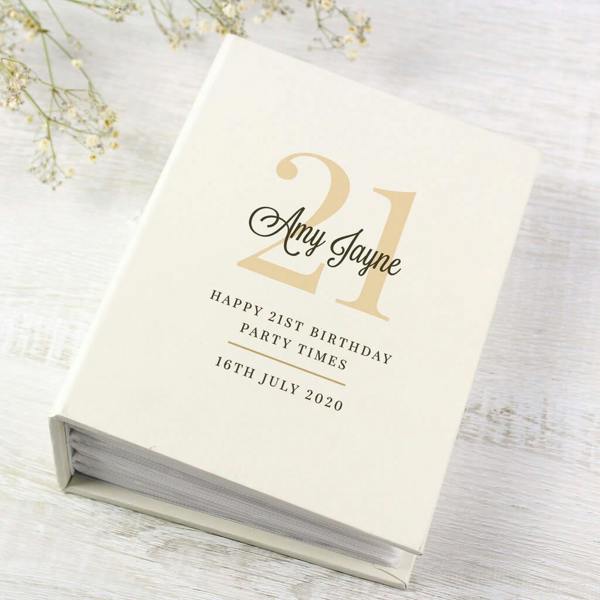 A personalised cream 21st birthday photo album printed with '21 Amy Jayne Happy 21st Birthday Party Times 16th July 2020' on a wooden surface next to white flowers.