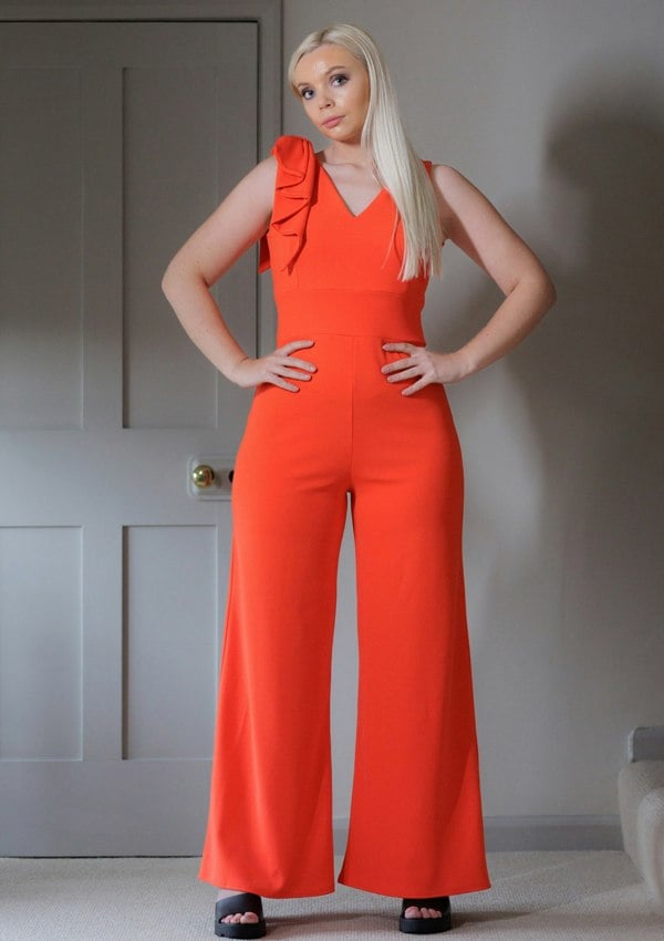 This is a young lady wearing a Flame red coloured V neck jumpsuit, with wide legs and a bow on one shoulder. She is wearing black open-toed sandals and has her hands on her hips.