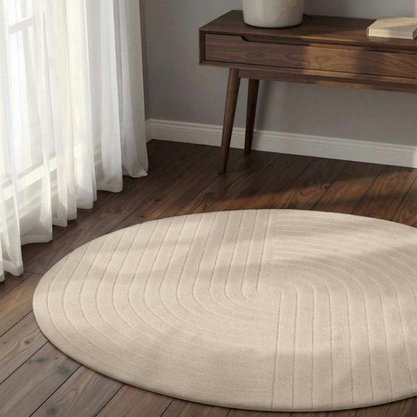 Round beige rug on a wooden floor with a wooden side table and white curtains.
